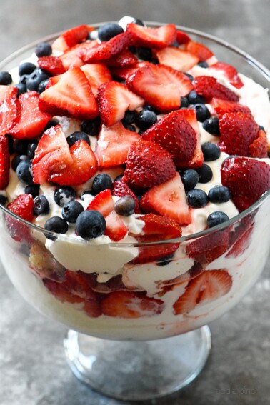 Glass trifle bowl holds a berry trifle dessert with layers of cream, vanilla cake, blueberries and strawberries.