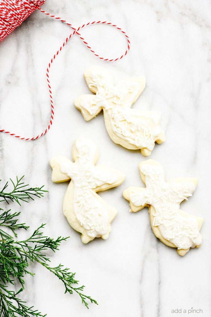 Cookies in shape of angels decorated with white icing and white sprinkles. Surrounded by greenery and red and white twine on a marble board.